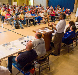 photo of a group of people sitting in folding chairs, government officials sit at a table in the front of the room.