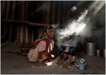 Woman sitting on the dirt floor of her home, filled with smoke from her cookstove