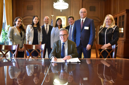 Acting Administrator Wheeler sits at a table holding a pen with a document in front of him as seven women and men stand behind him  in a large room