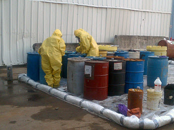 two people in yellow haz-mat body suits surrounded by many 55-gallon drums and buckets