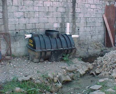 The photo shows a septic tank sitting above ground next to a building and the hole in the ground into which it will be installed.