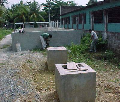 The photo shows workers beside the walls of the concrete rectangular sand filter with the two square concrete access ports in the foreground.  The outside wall of the school is shown directly to the right of the sand filter.