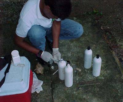 The photo shows a person wearing gloves kneeling on the ground beside a cooler and several sample containers.