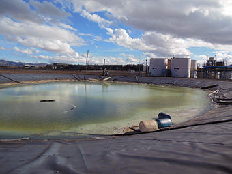 Plástico negro líneas de material laminado una depresión llena de líquido verde con 3 barriles de plástico al borde del agua, blancos tanques de almacenamiento de metal en el fondo.